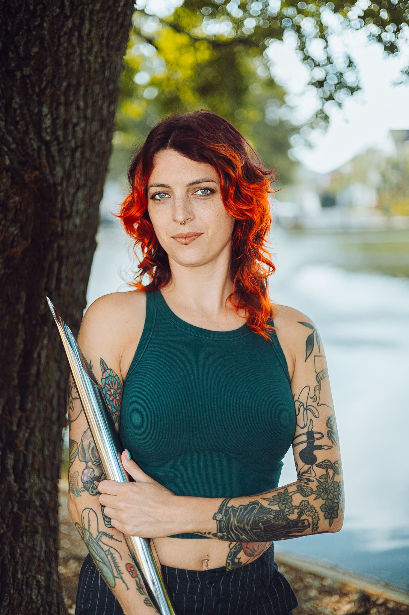 Portrait of a tattooed woman with bright orange hair, standing by a tree outdoors and holding a metallic rod/lantern.