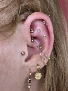 Close-up of an ear with multiple gold earrings and chains, including small stud pegs on the lobe and a hanging chain jewelry piece along the ear canal.