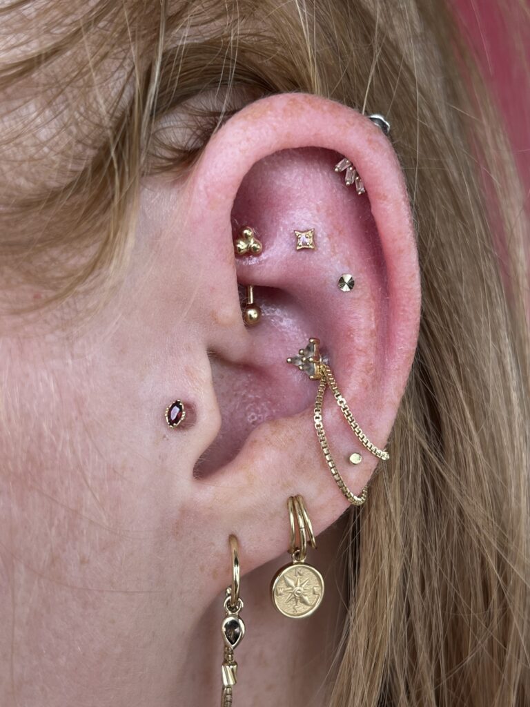 IMG_6064 | Red Baron Ink Tattoo & Piercing Close-up of an ear with multiple gold earrings and chains, including small stud pegs on the lobe and a hanging chain jewelry piece along the ear canal.